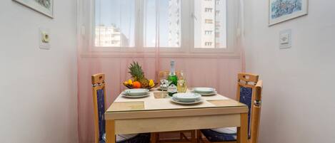 Mesa de jantar posta para quatro pessoas, com cadeiras, uma janela com cortinas leves e obras de arte nas paredes.