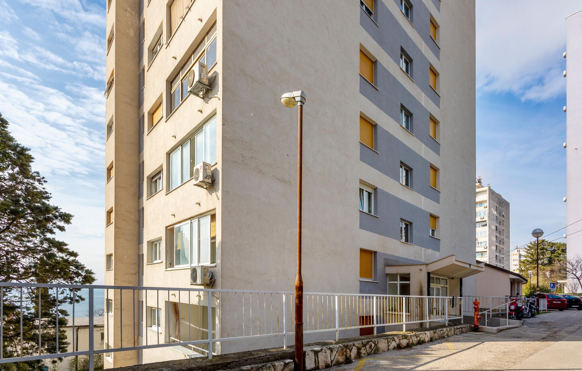 Un edificio residenziale a più piani con balconi, un ingresso con cancello e un’area parcheggio con auto e motociclette.