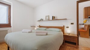 A bedroom with a bed, bedside tables, a shelf with books and a picture, a window, and a view of a kitchen.