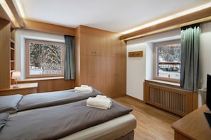 A hotel room with two beds, wooden wardrobe, a radiator, and large windows with curtains.
