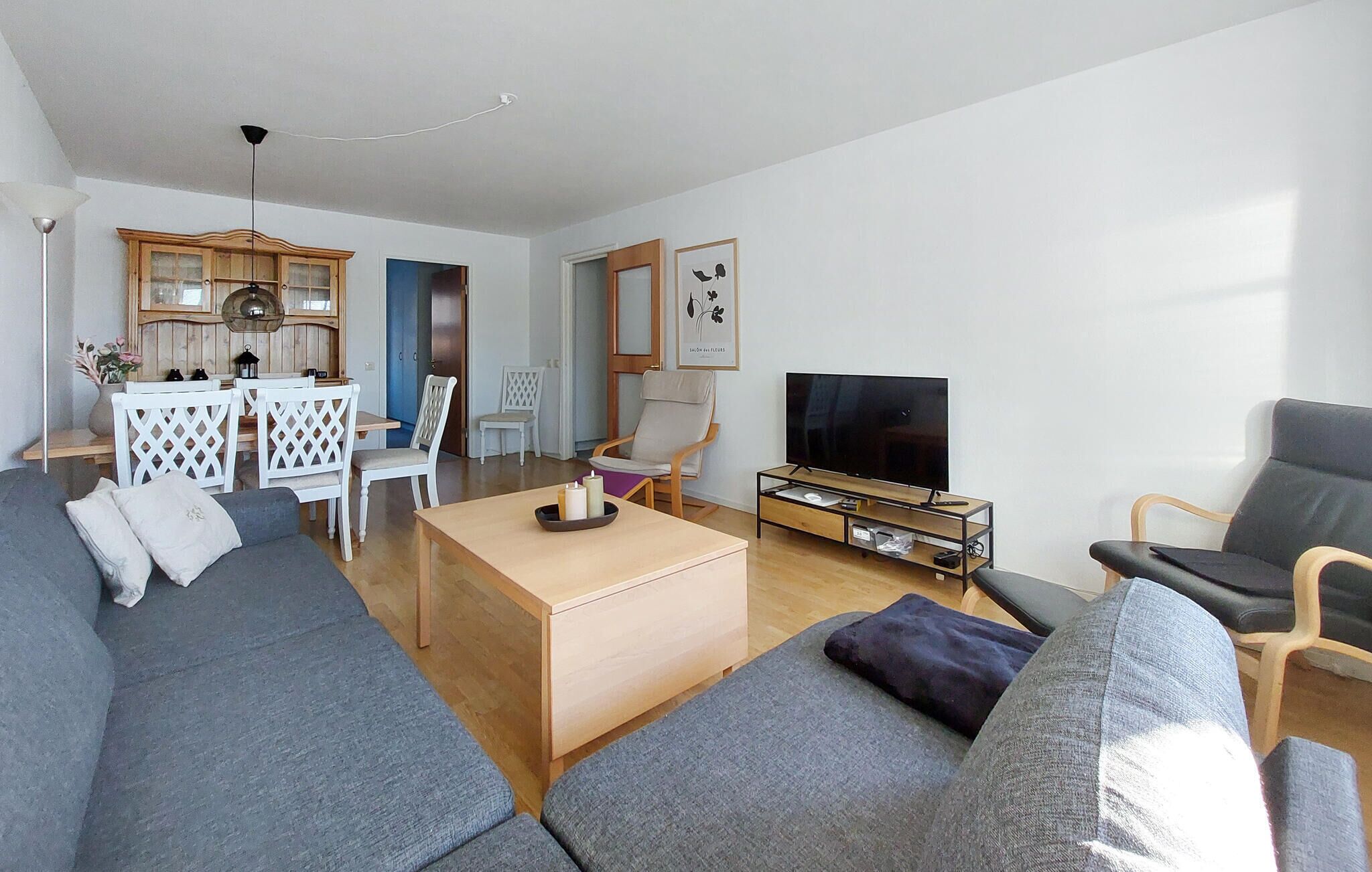 A modern living room with a grey sofa, a wooden coffee table, a TV on a stand, and a dining area with white chairs and a wooden cabinet.