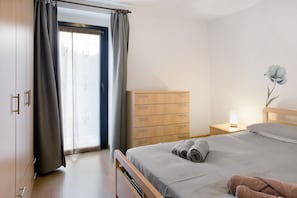 Un dormitorio con cama, cómoda de madera, mesita de noche con lámpara y ventana con cortinas.