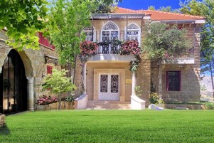 Un edificio de piedra con techo de tejas rojas, una entrada central con puerta blanca y un pequeño balcón lleno de flores.