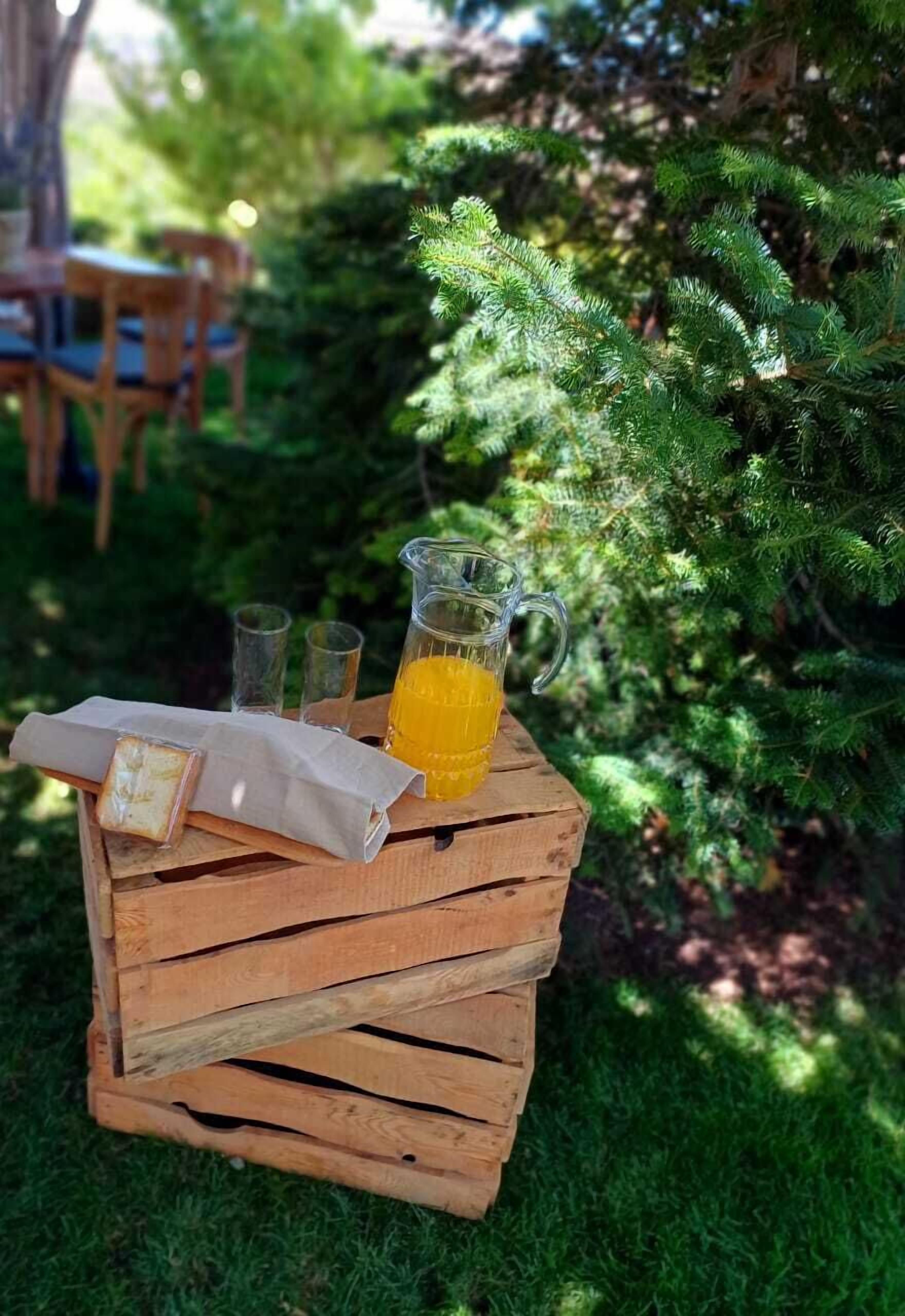 Una caja de madera con una jarra de vidrio de jugo de naranja, dos vasos y una servilleta sobre una zona verde al aire libre.