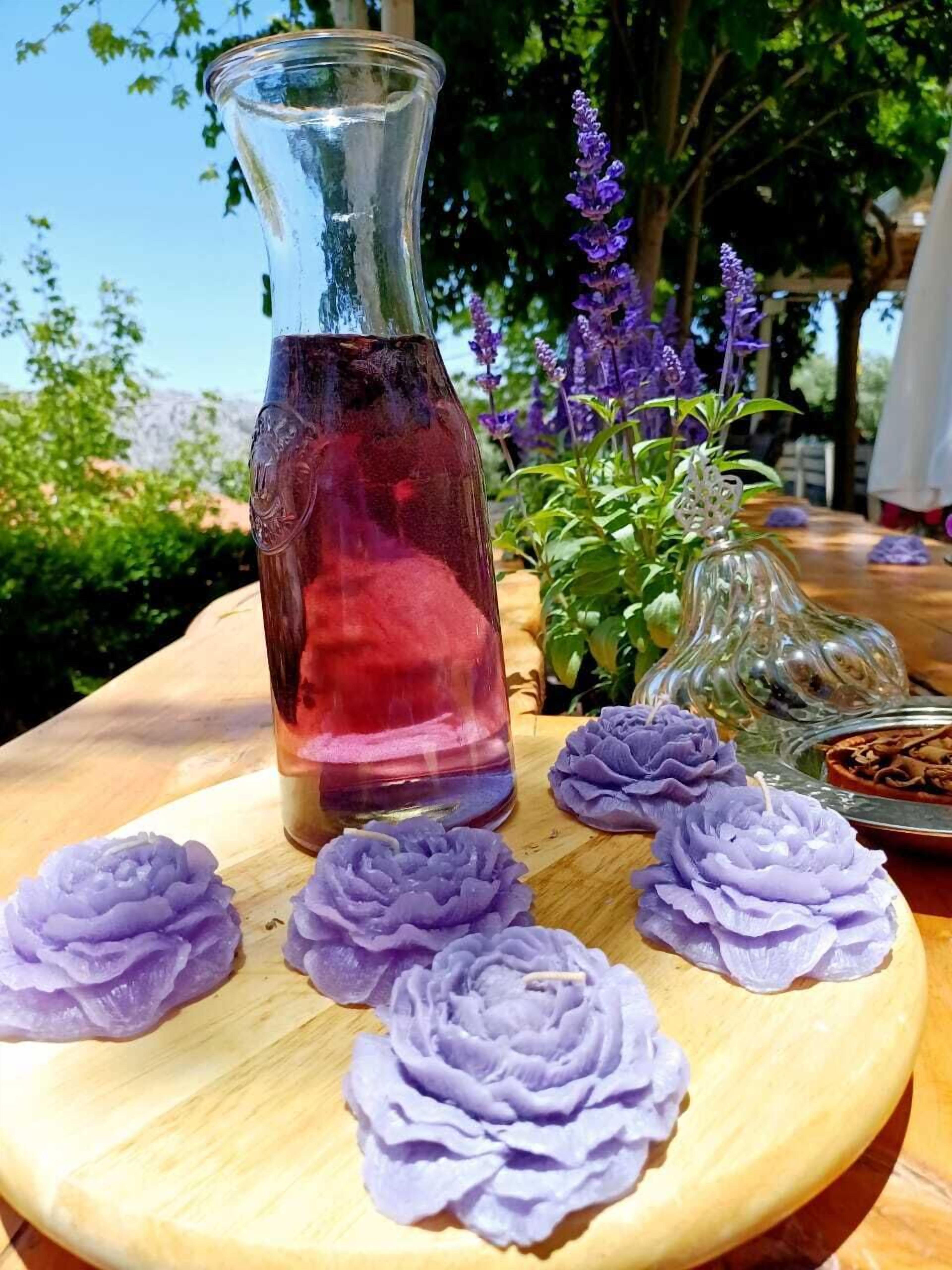 A clear glass vase with a red liquid, surrounded by purple flowers on a wooden table outdoors.