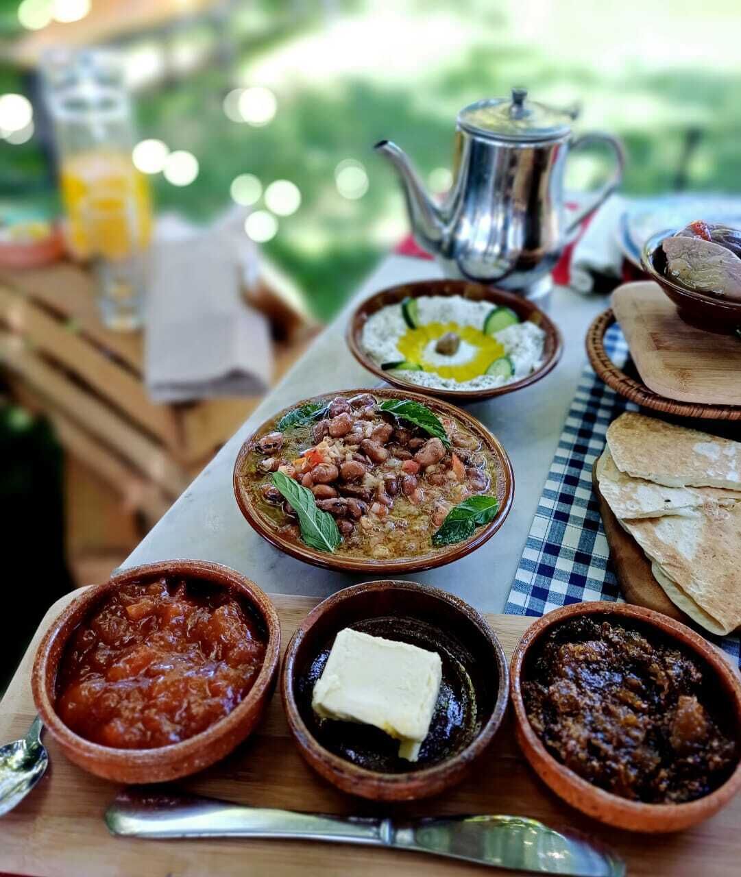 Una mesa con varios platos, entre ellos un tazón de frijoles, un tazón de salsa de tomate y un tazón de carne. También hay una tetera y un vaso de jugo.