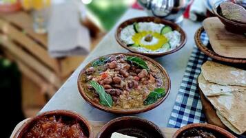 Una mesa con varios platos, entre ellos un tazón de frijoles, un tazón de salsa de tomate y un tazón de carne. También hay una tetera y un vaso de jugo.