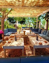 Un patio exterior con zona de estar cubierta, mesas de madera y una fuente de piedra.