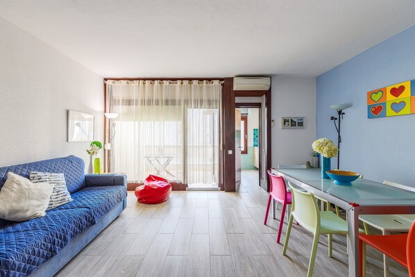 Un salon avec un canapé, une table à manger, des chaises colorées et une vue sur l’extérieur, visible à travers une porte.
