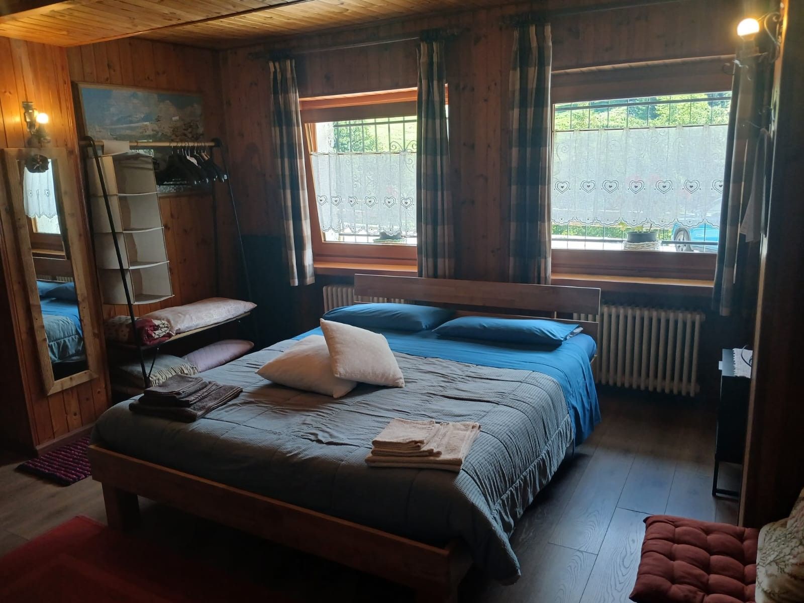 Una stanza in legno con un letto, una panca, un termosifone e una finestra con tende.