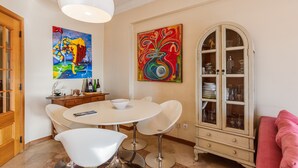 Un comedor con una mesa redonda, sillas blancas, un mueble con puertas de vidrio y dos cuadros en la pared.