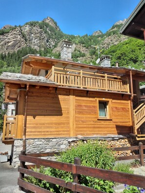 A wooden cabin with a balcony, stone foundation, and a mountainous backdrop.