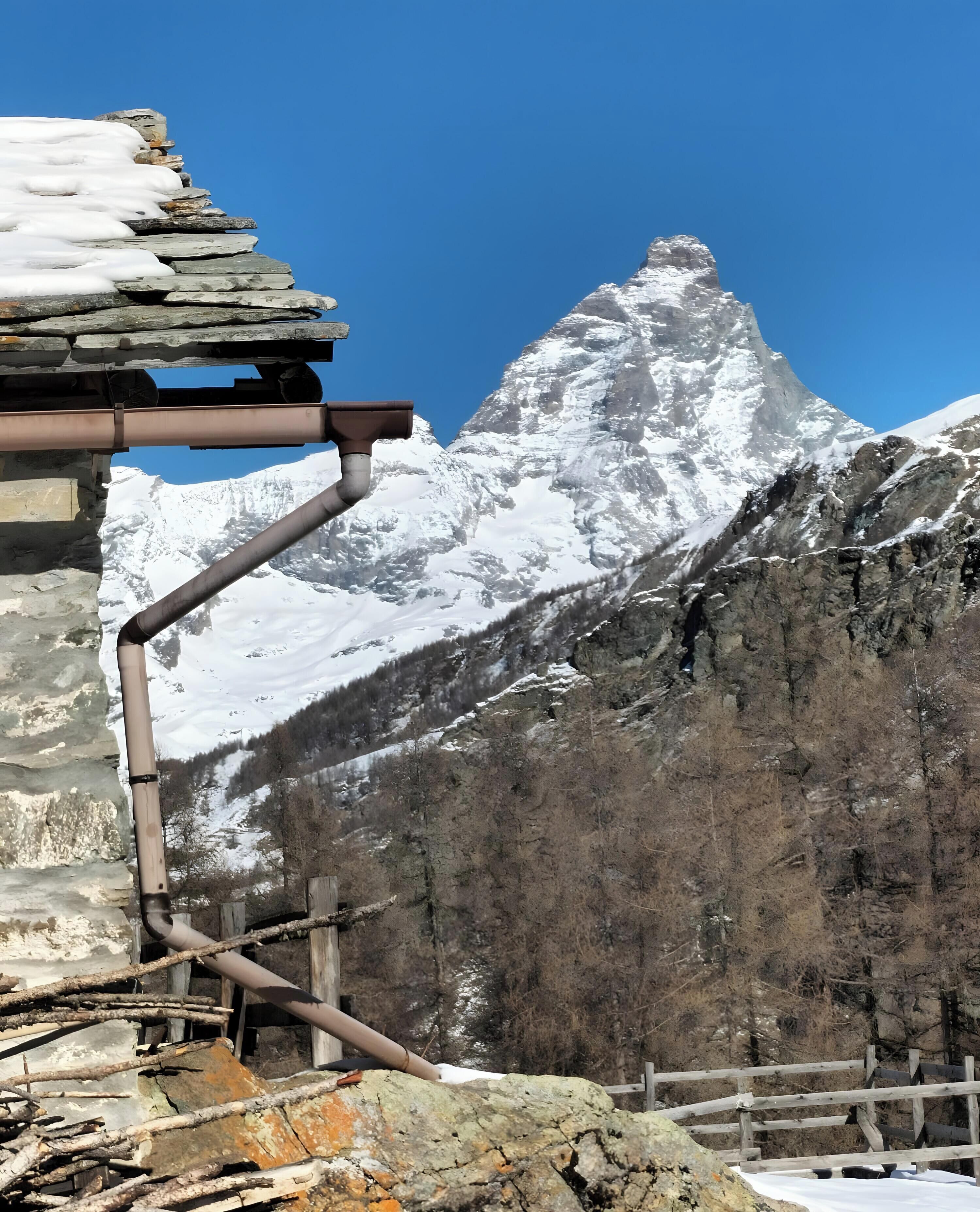 A mountain with a snow-covered peak, a wooden fence, and a stone building with a metal pipe.