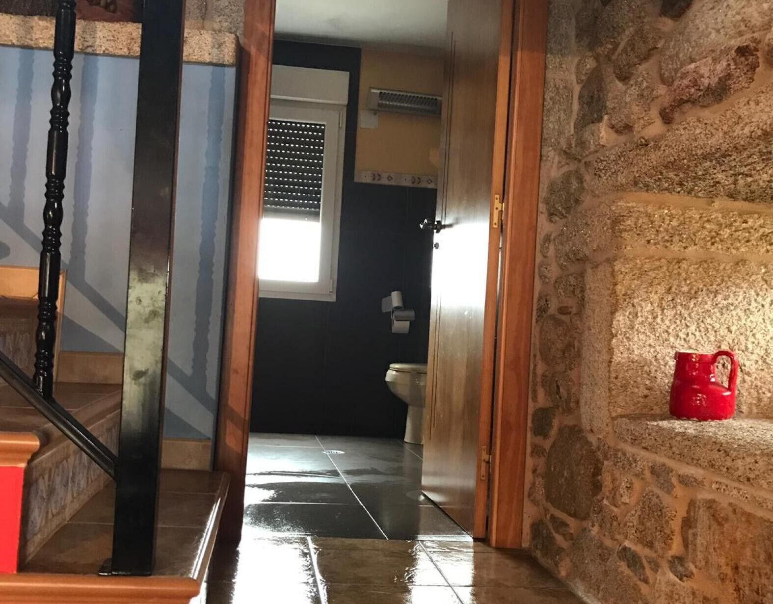 Een hal met een trap, een badkamer met toilet en raam, en een stenen muur.