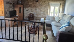 A stone-walled room with a wooden ceiling, a television on a wooden stand, a leather armchair, a sofa, and a glass coffee table. - Country House 'Fidalgo' with Mountain View, Private Terrace and Wi-Fi (Vimianzo)