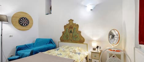 A bedroom with a bed, a chair, a bedside table with a lamp, a wall clock, and a fan.