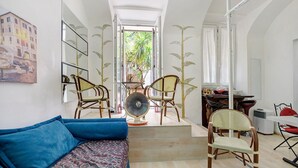 A narrow hallway with a sofa, chairs, a fan, and a staircase leading to an outdoor area with a view of palm trees.