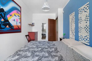 Une chambre à coucher avec un lit, une chaise rouge, un bureau, une lampe, un tableau et une porte qui donne sur une autre pièce.