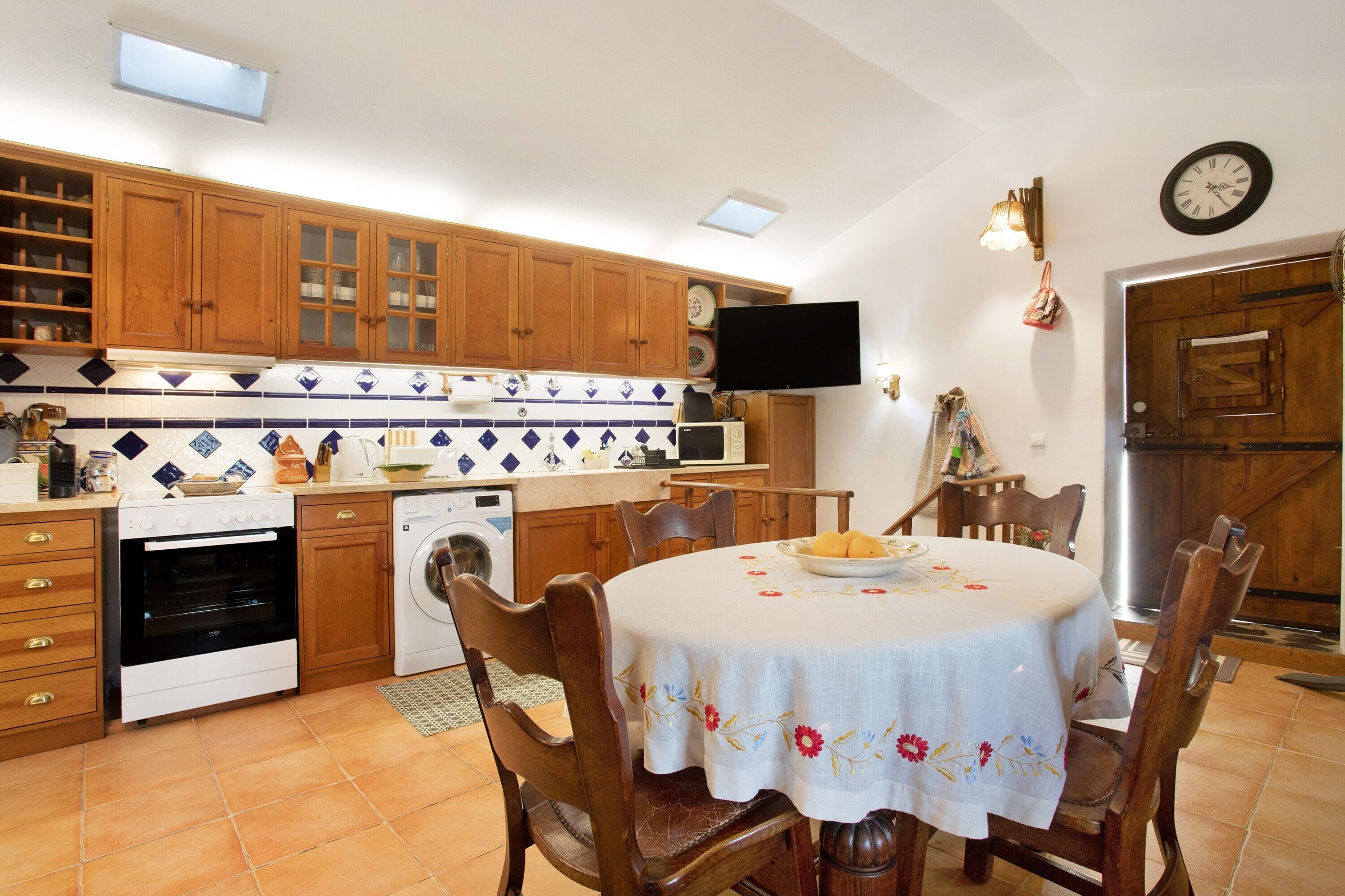 Una cocina con armarios de madera, una mesa de comedor con sillas, una lavadora y un televisor.