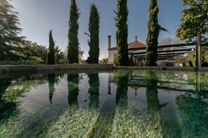 Un étang aux eaux claires où se reflètent de grands arbres et un bâtiment surmonté d’un toit rouge.