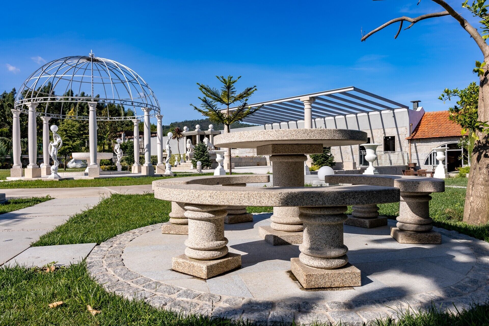 Un jardin agrémenté d’une fontaine en pierre, d’une structure surmontée d’une coupole de verre et d’un bâtiment au toit rouge.