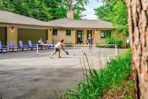 An outdoor tennis court with a net, surrounded by lounge chairs and a building with a stone chimney. - Woodsy Waterfront Retreat on Gorgeous Balsam Lake (Balsam Lake)