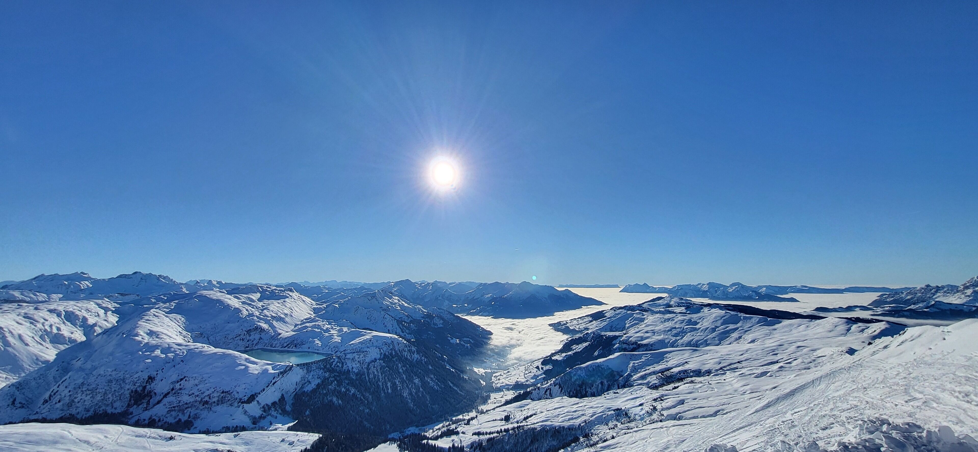 Vista panorámica de un paisaje montañoso cubierto de nieve, con cielo despejado y el sol brillando intensamente.