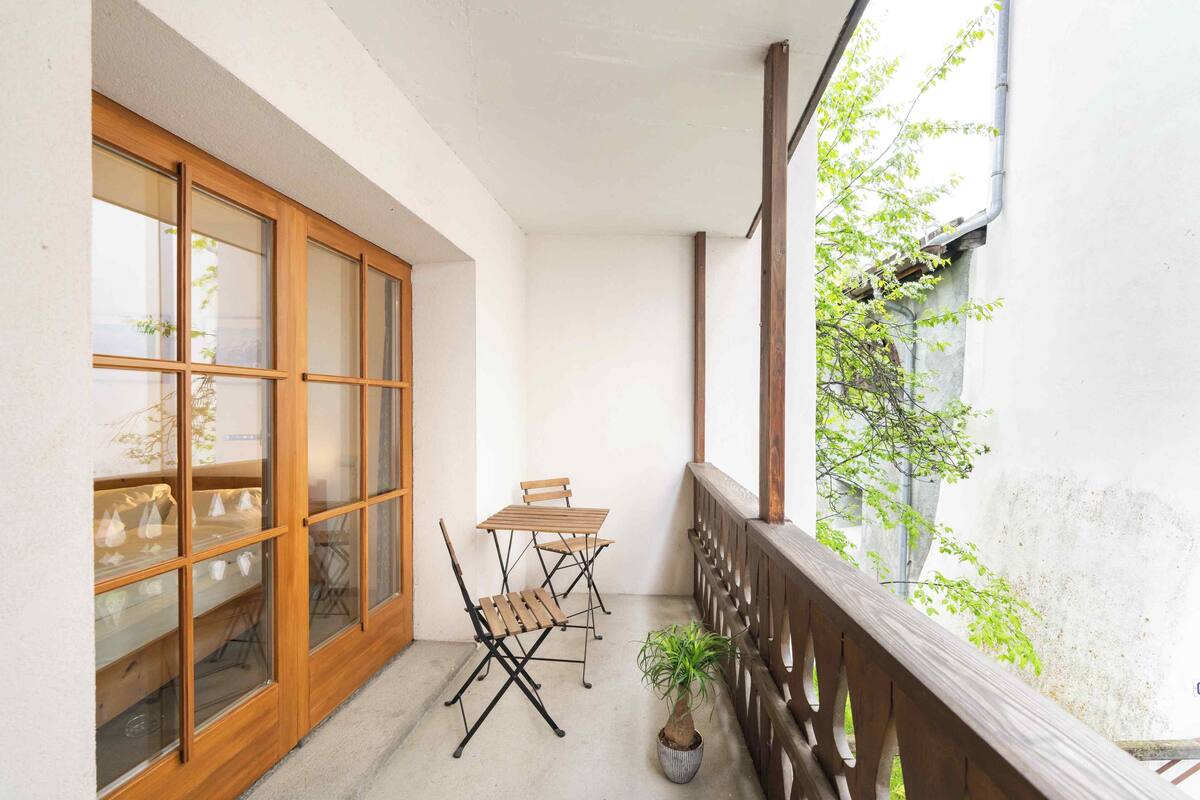 A balcony with a wooden table and chairs, a potted plant, and a view of a building with large windows and a tree.