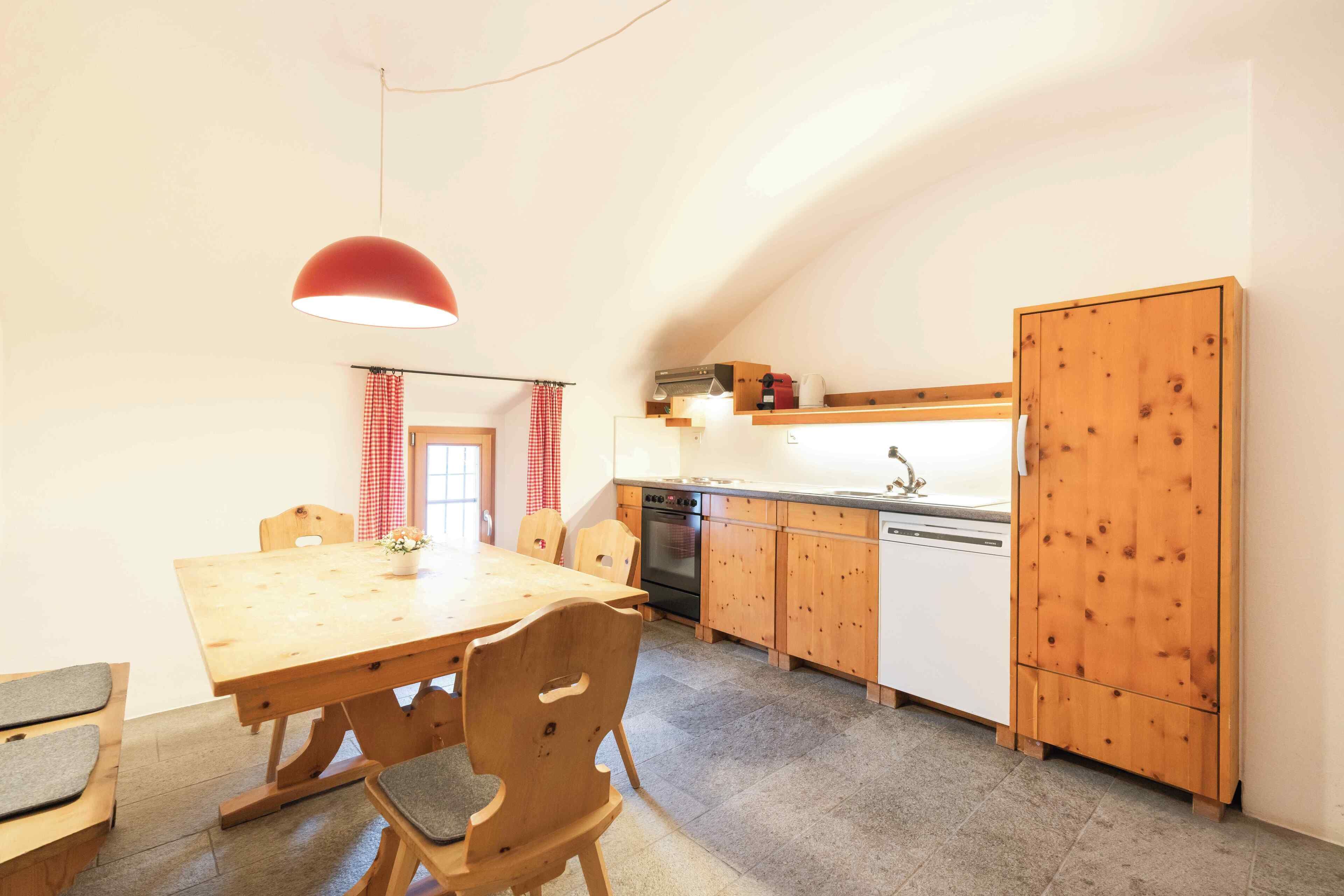 Een eetruimte met een houten tafel en stoelen, een rode hanglamp, en een keuken met houten kastjes en een vaatwasser.