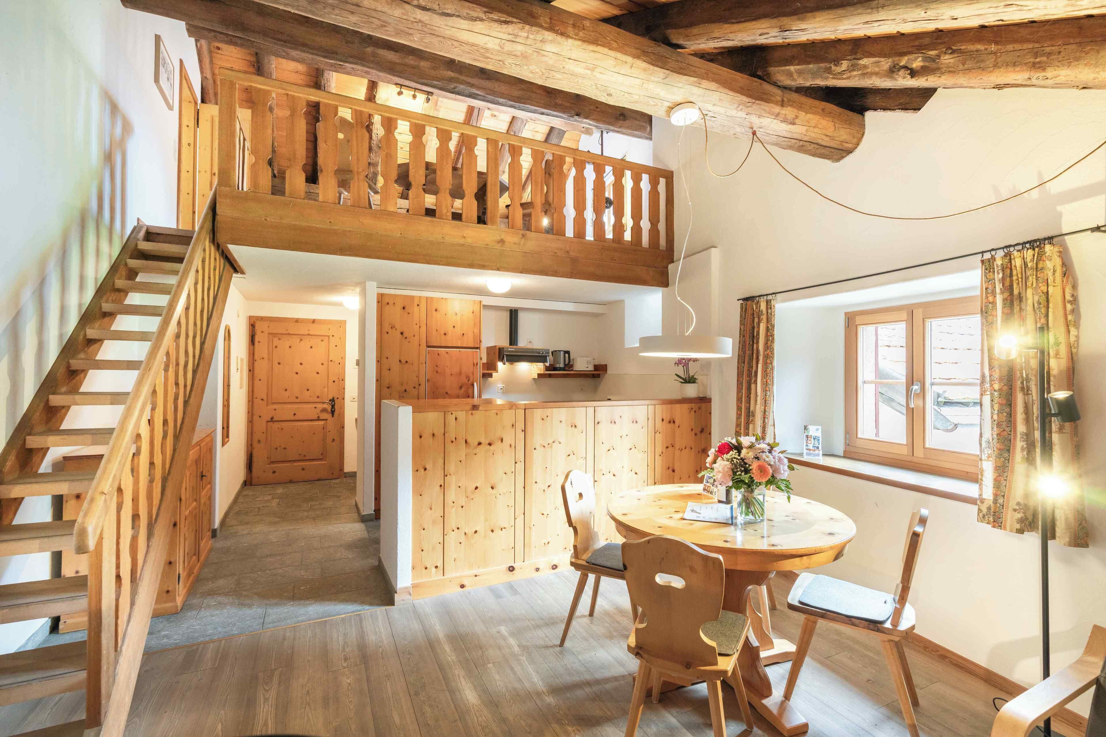 A wooden staircase leading to a loft bedroom with a balcony, a kitchen with wooden cabinets, and a dining area with a round table and chairs.