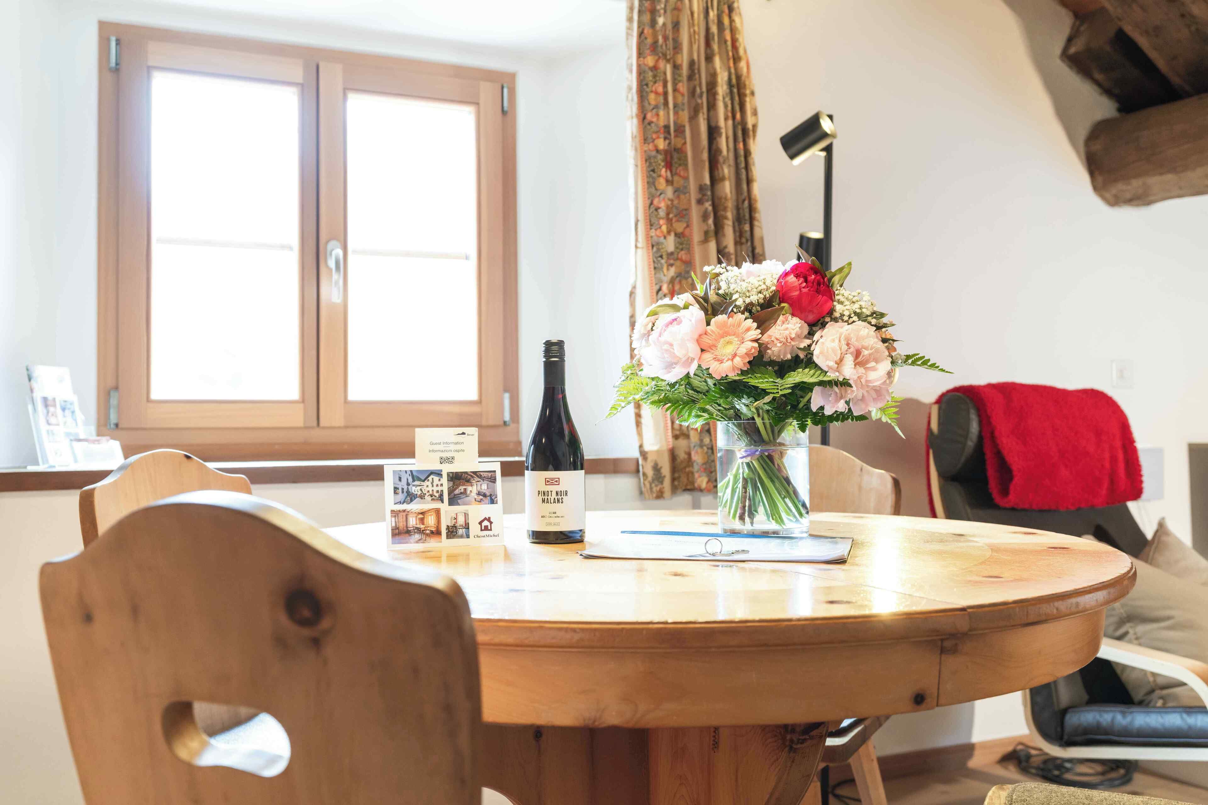 Una mesa de comedor con una botella de vino tinto Pinot Noir Malans, un ramo de flores y una tarjeta de información para huéspedes.