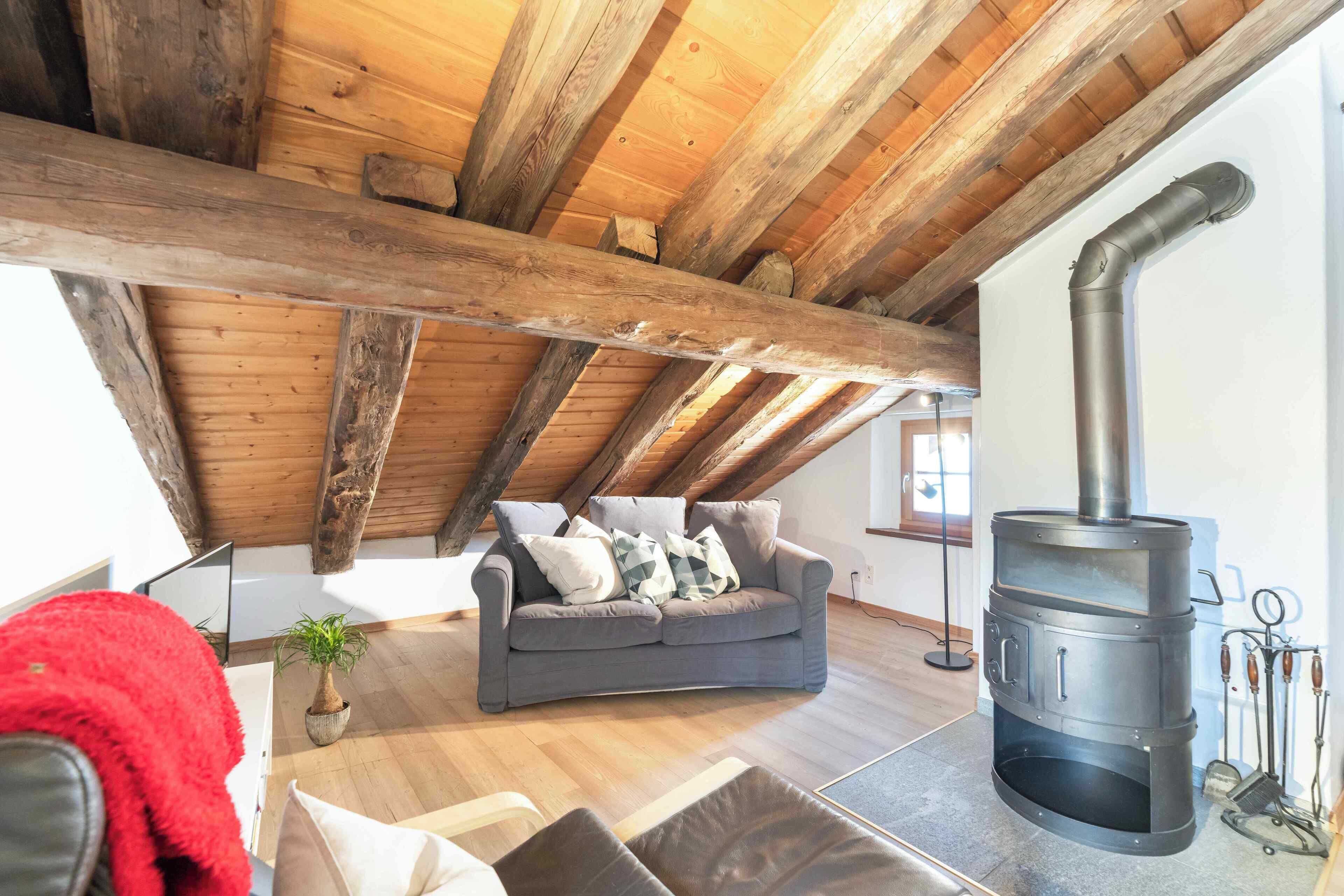 Une chambre à coucher confortable, aménagée en mezzanine, avec un plafond en pente, un poêle à bois encastré, un canapé gris et une couverture rouge sur le lit.