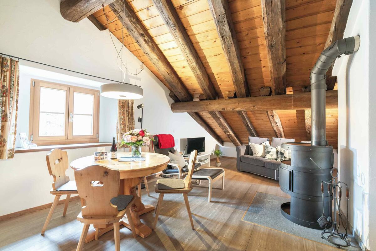 A cozy loft with a wooden ceiling, a dining area with a round table and chairs, a sofa, and a wood-burning stove.