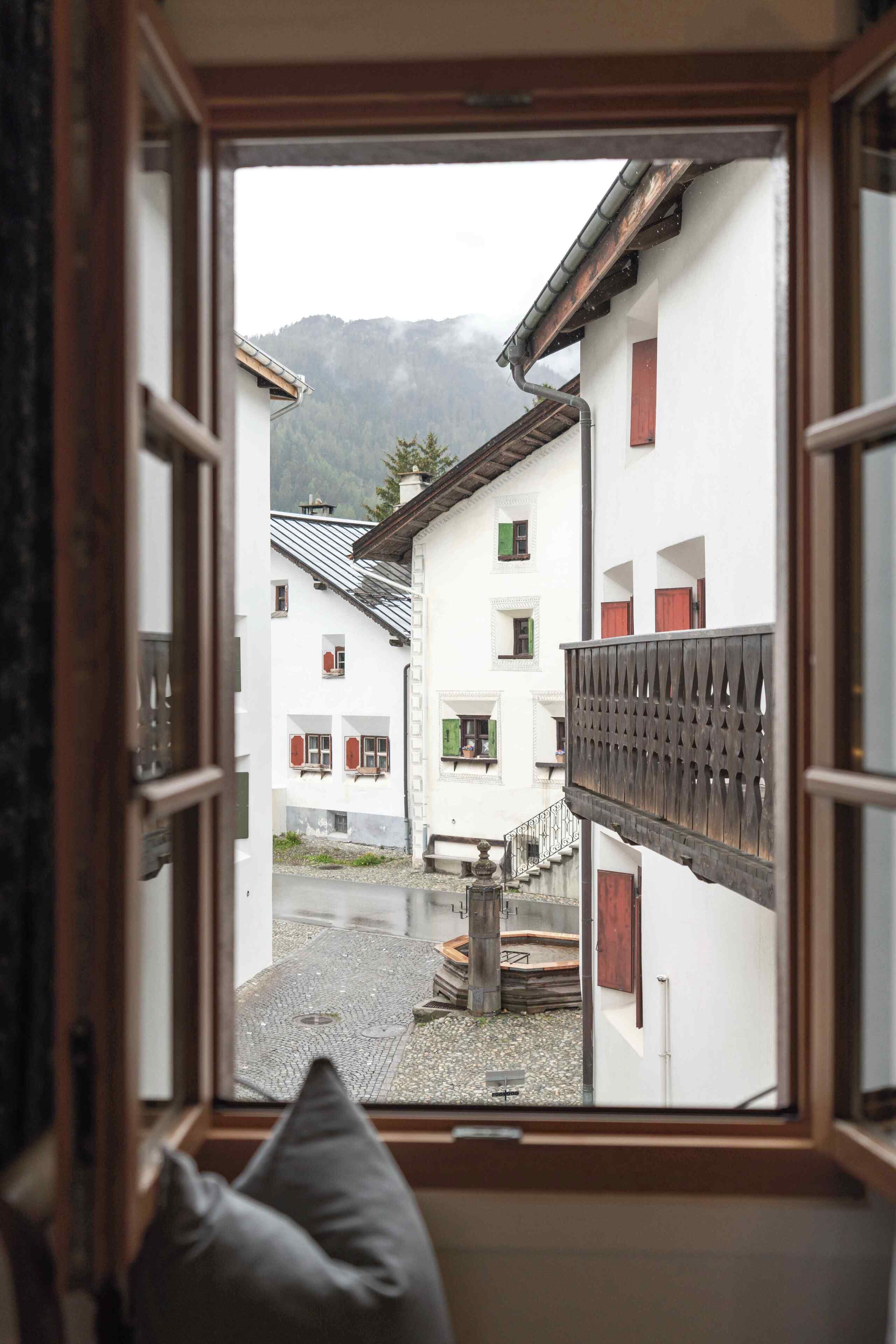 A view through an open window of a courtyard with buildings, a wooden fence, and a mountain in the background.