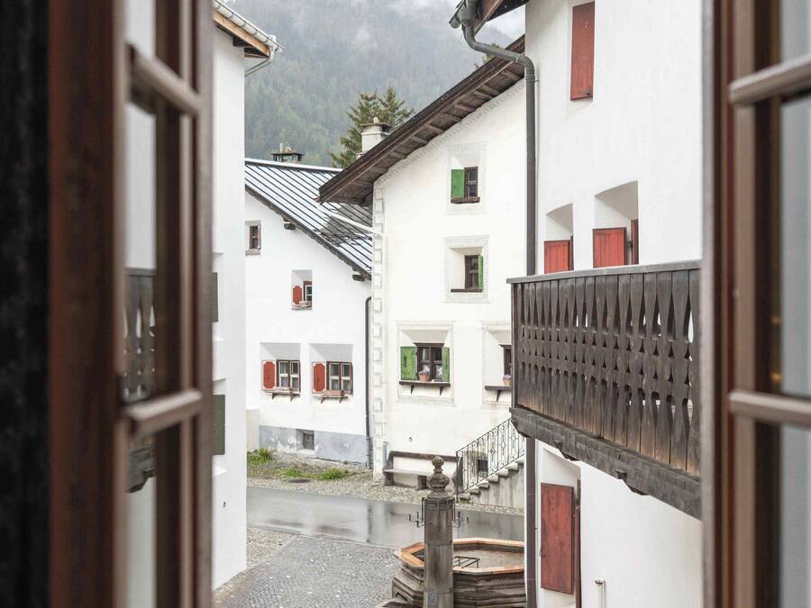 A view through an open window of a courtyard with buildings, a wooden fence, and a mountain in the background.