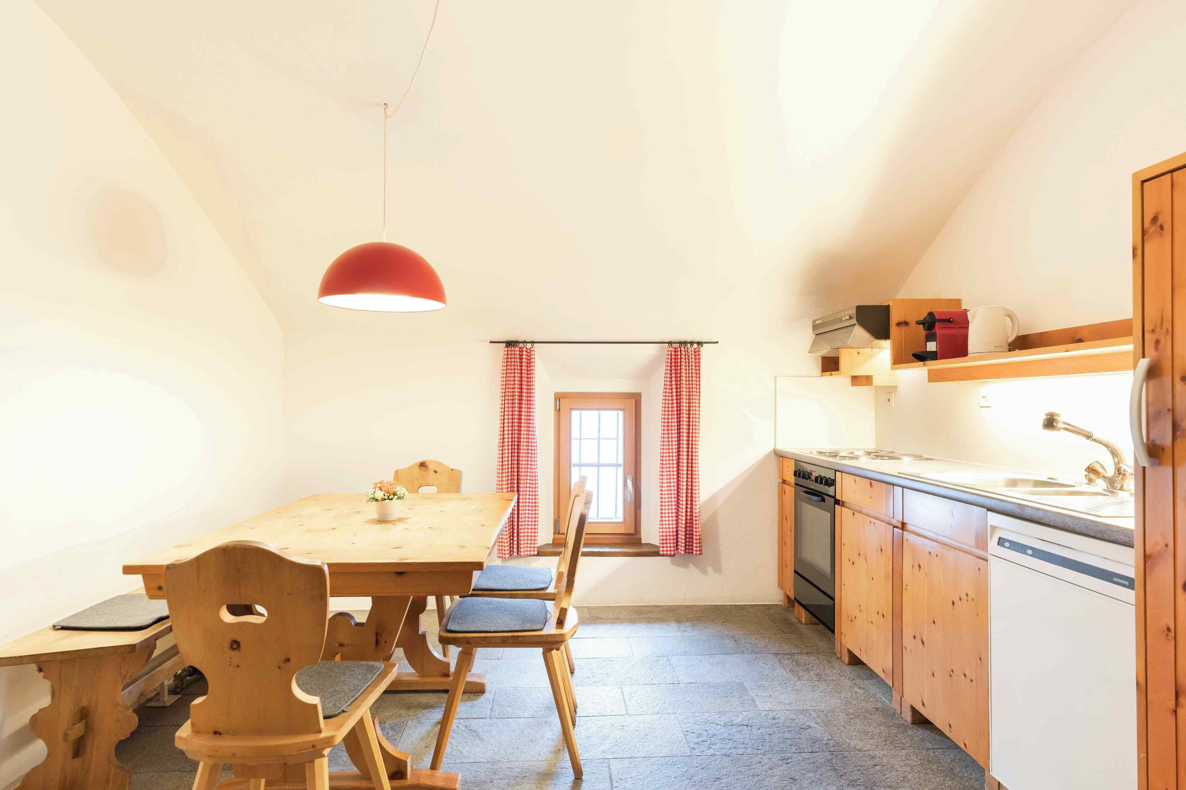 Een keuken met houten kastjes, een houten eettafel en stoelen. Boven de tafel hangt een rode hanglamp.