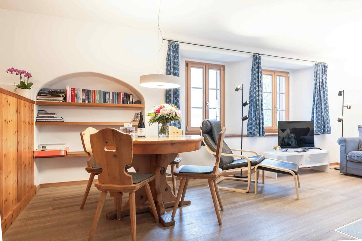 A cozy living room with a wooden dining table, chairs, a bookshelf, a TV, and a window with curtains.