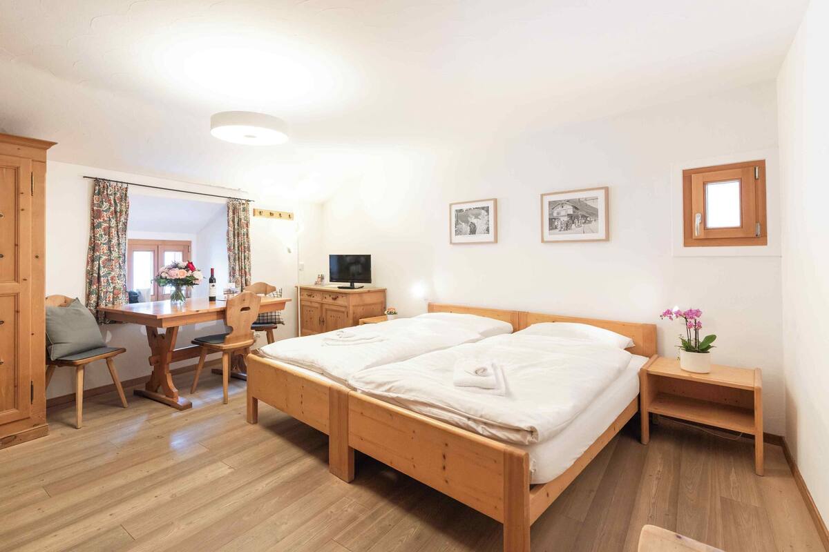 A room with a wooden bed, a desk with a chair, a small table, and a TV.