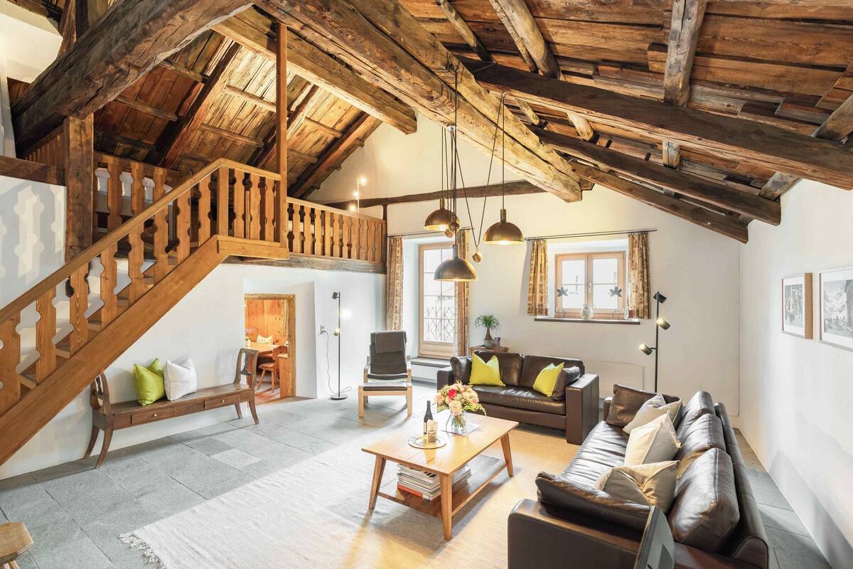 A spacious living area with a wooden staircase, a leather sofa, a coffee table, and a dining area with a chair and table.