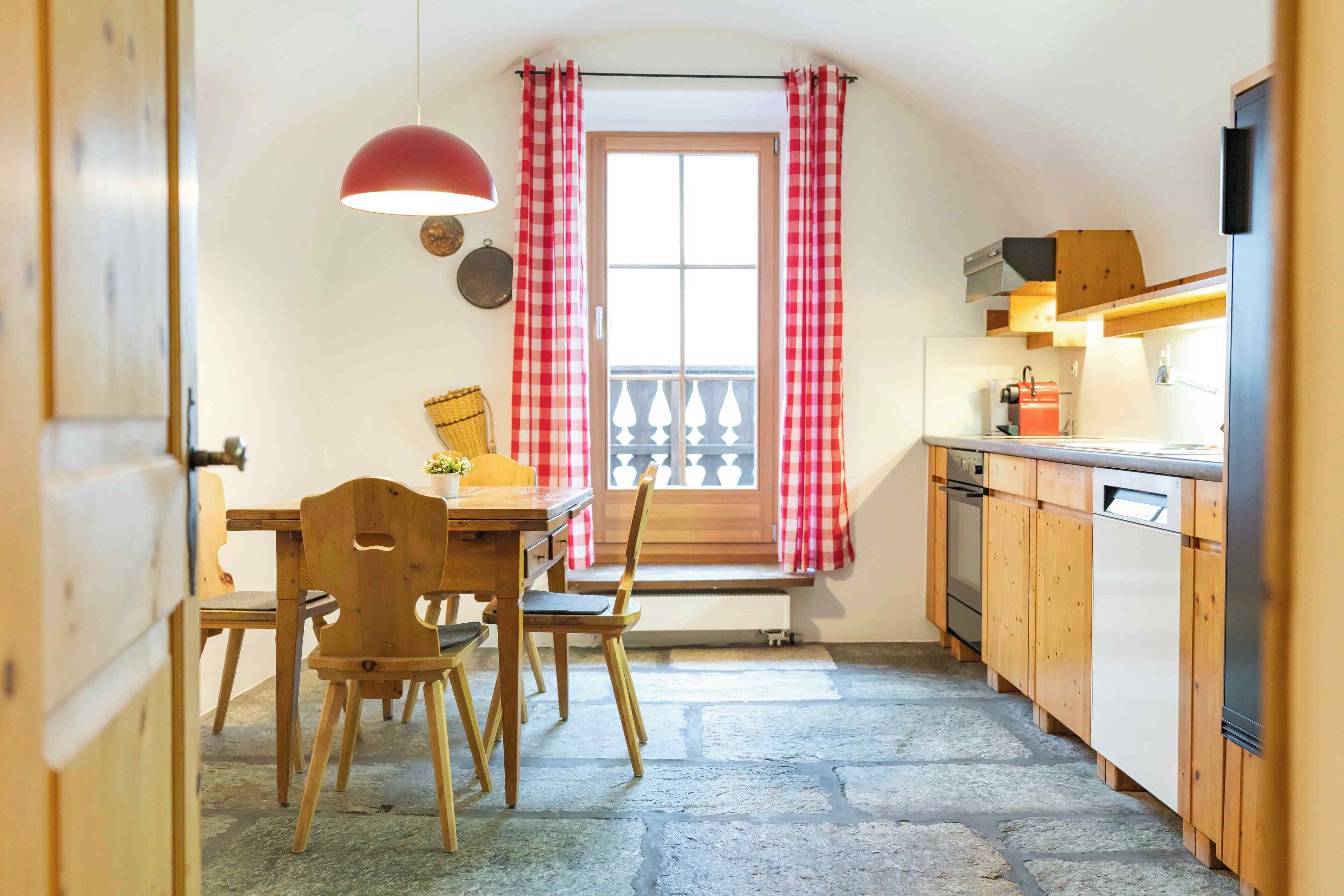 Une cuisine avec des meubles de rangement en bois, une table à manger avec des chaises, un rideau à carreaux rouges et blancs, et une fenêtre donnant sur un balcon.