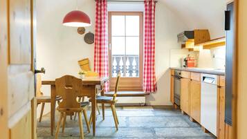 Une cuisine avec des meubles de rangement en bois, une table à manger avec des chaises, un rideau à carreaux rouges et blancs, et une fenêtre donnant sur un balcon.