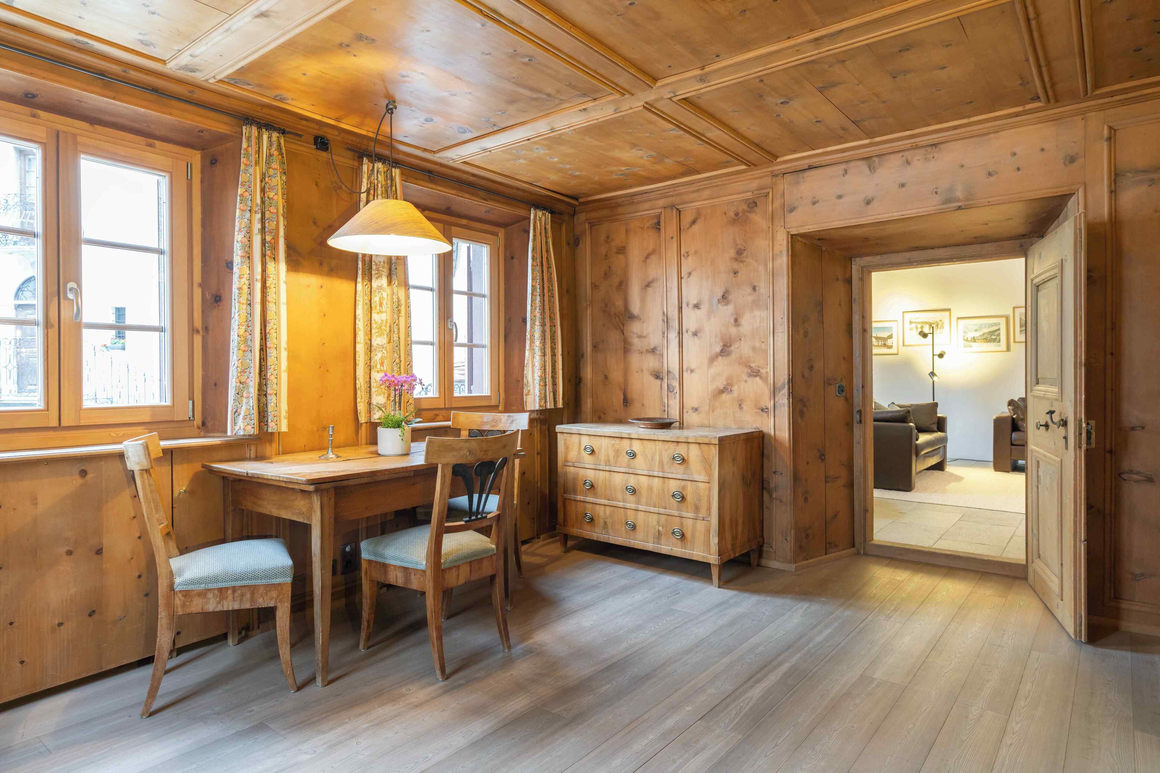 A wooden interior with a dining table, chairs, a dresser, and a door leading to another room.