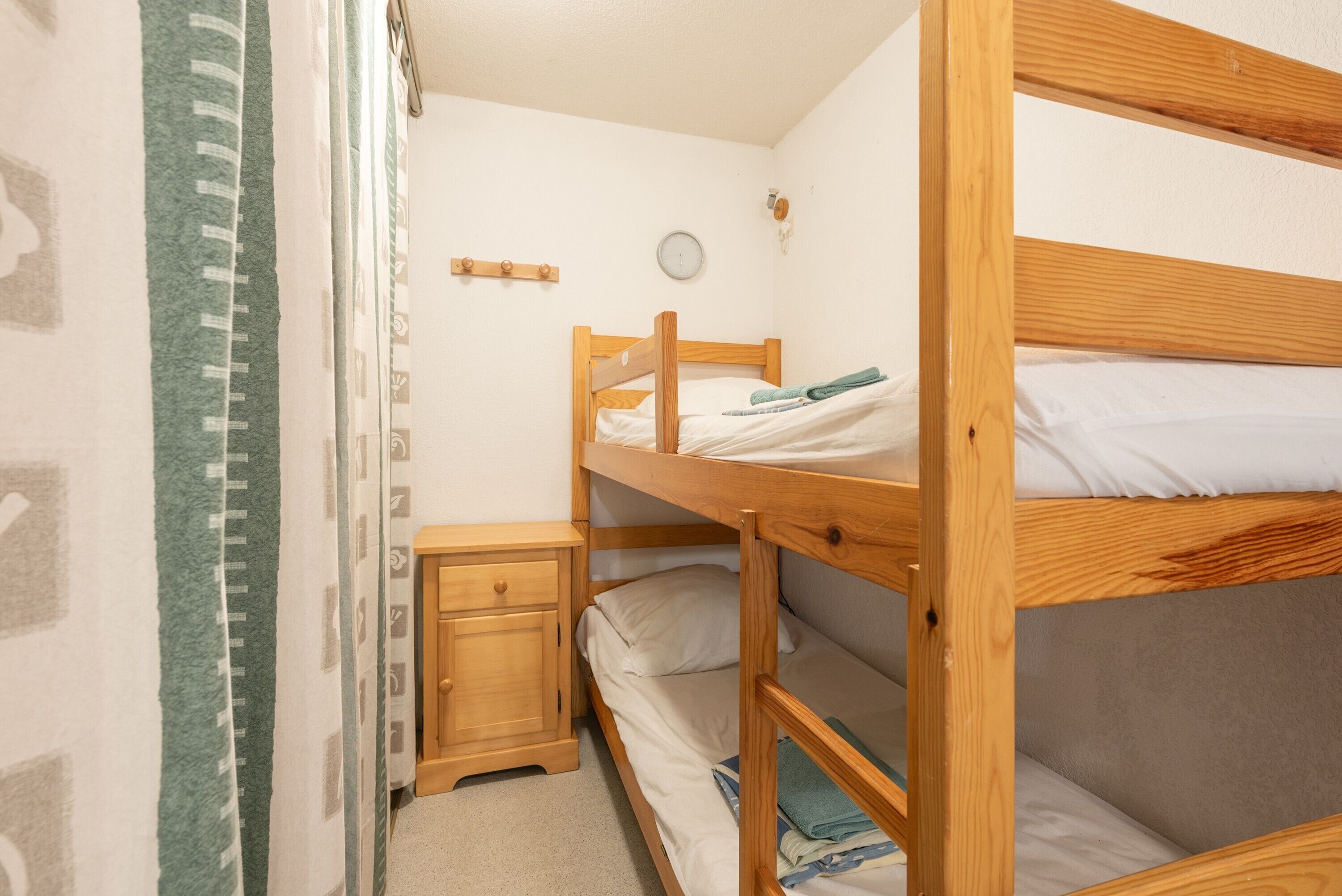Habitació amb lliteres, un panell de capçal de fusta, una petita taula de nit, un rellotge de paret i una cortina.