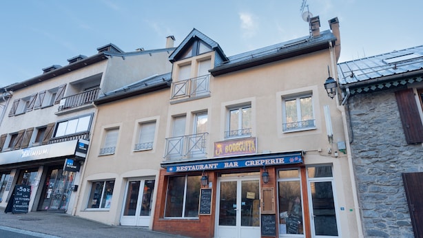A building with a restaurant named 'BODEGUITA' on the ground floor, a 'BRIMIUM' store on the left, and a stone wall on the right. - Cosy Stay in Barèges – Mountain Views, Wi-Fi, Free Parking, Near Ski Slopes (Barèges)