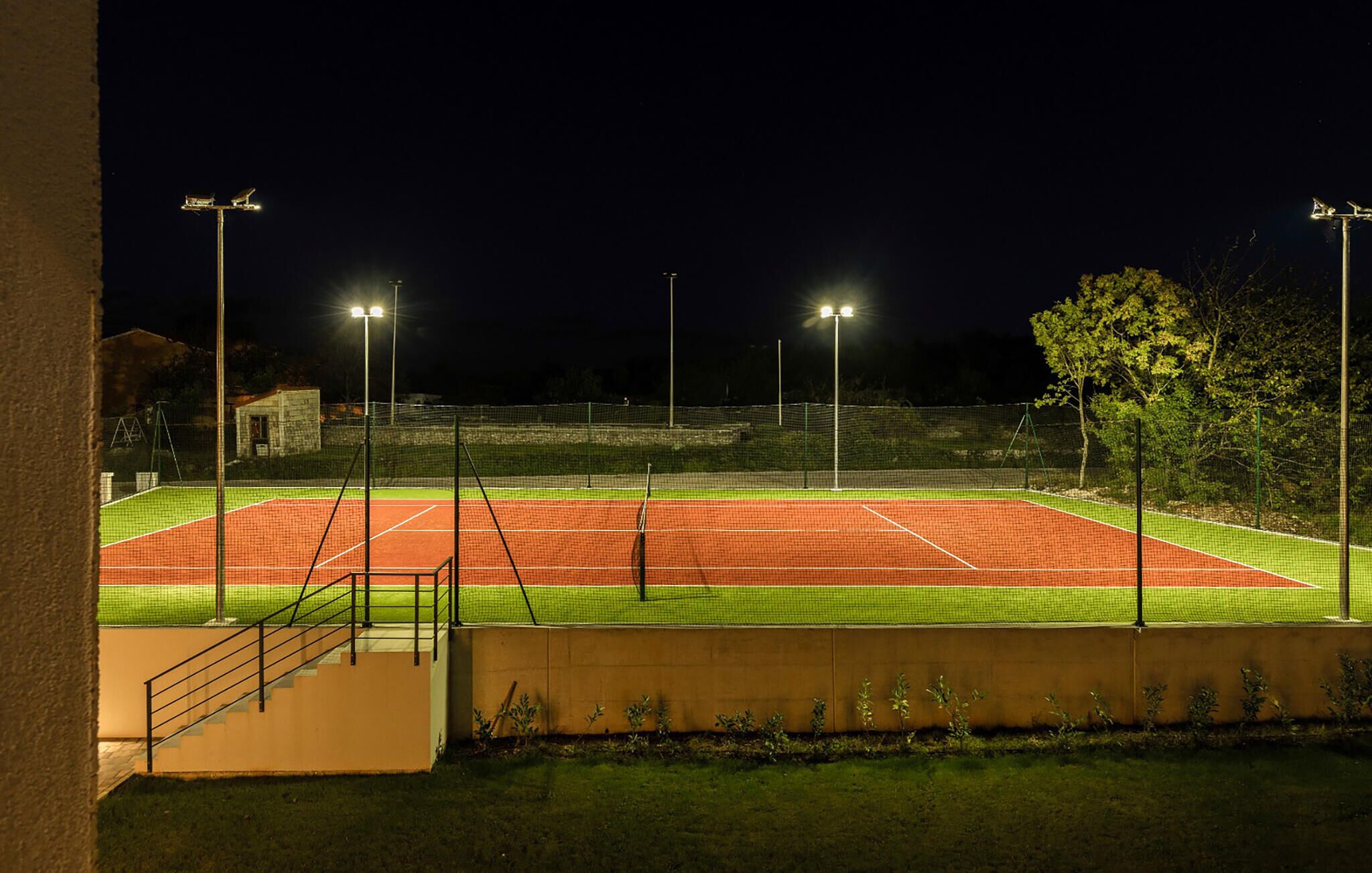 Tenniskenttä yöllä, jossa on valonheittimet, verkko ja sitä ympäröivä aita.