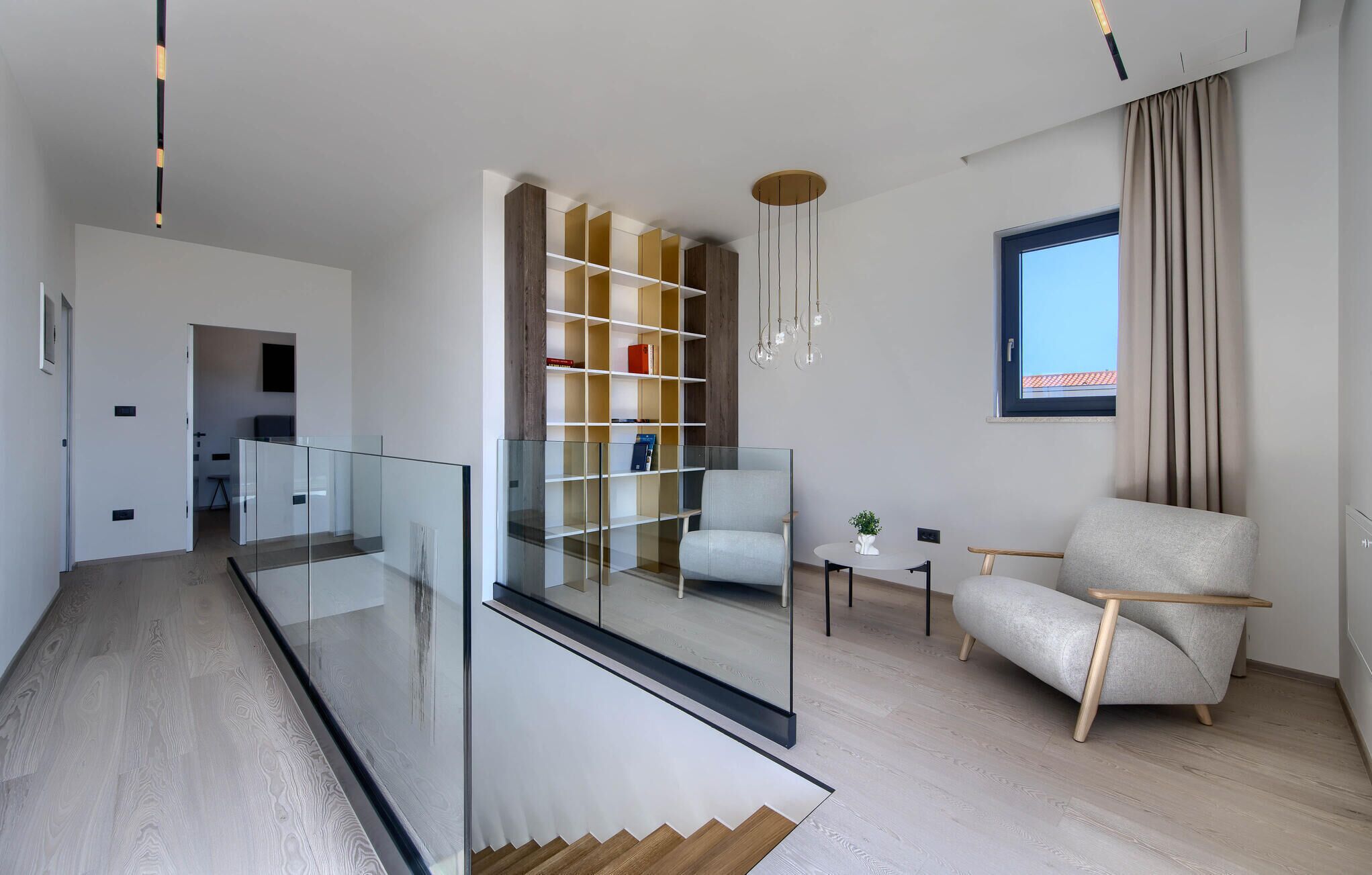 Una sala de estar moderna con una escalera de cristal, suelo de madera, una estantería, un área de estar y un ventanal con cortinas.