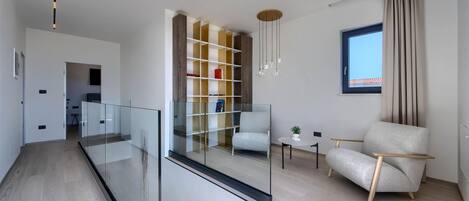 Una sala de estar moderna con una escalera de cristal, suelo de madera, una estantería, un área de estar y un ventanal con cortinas.