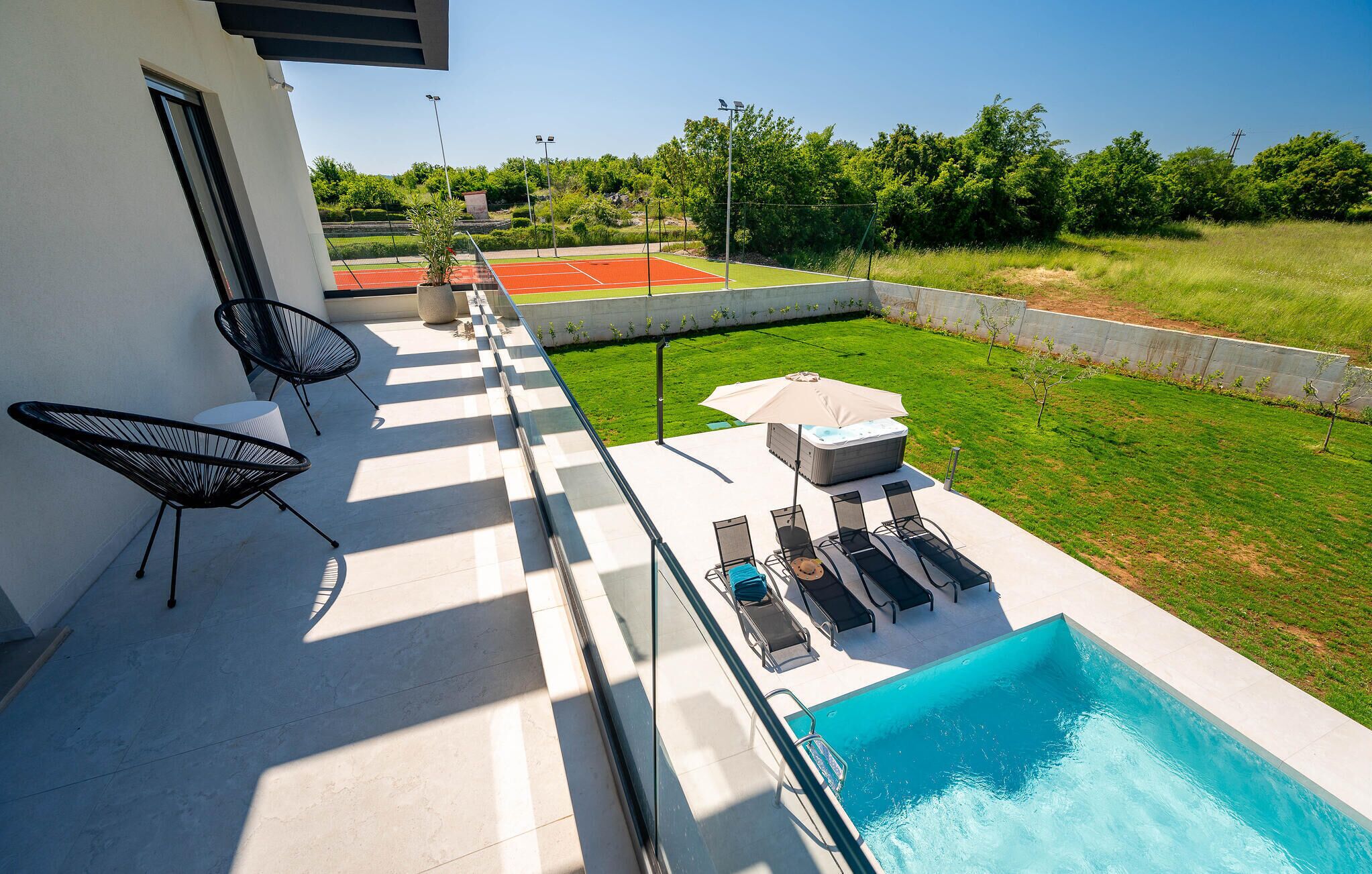 A modern outdoor pool area with a hot tub, lounge chairs, and a tennis court in the background.