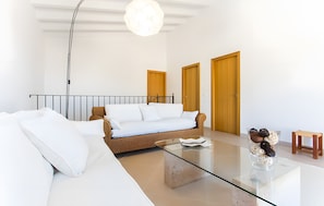 Een moderne woonkamer met een bank, een glazen salontafel en een rieten stoel. Er zijn twee houten deuren en een klein houten krukje.
