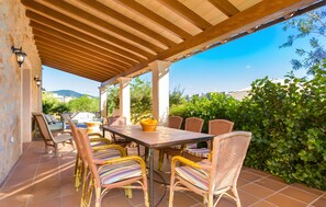 An outdoor patio with a wooden pergola, wicker chairs, a table, and a view of greenery and buildings in the background.